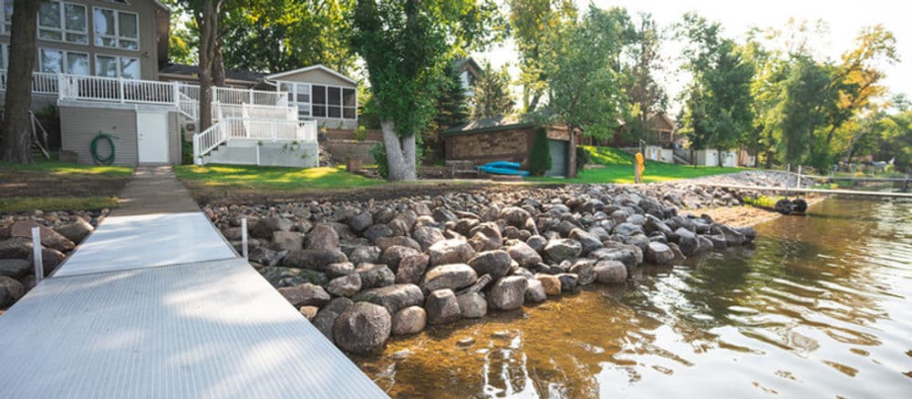 Lakeside view with a dock, rocky shore, houses, and greenery on a sunny day.