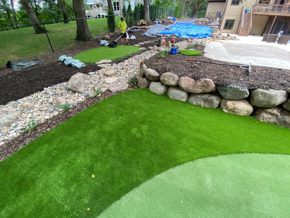 Lush green artificial turf installation near stone wall in landscaped yard setting.