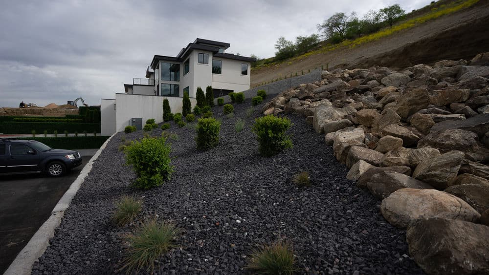 Modern house with landscaping featuring rocks and greenery on a hillside. Cloudy sky above.