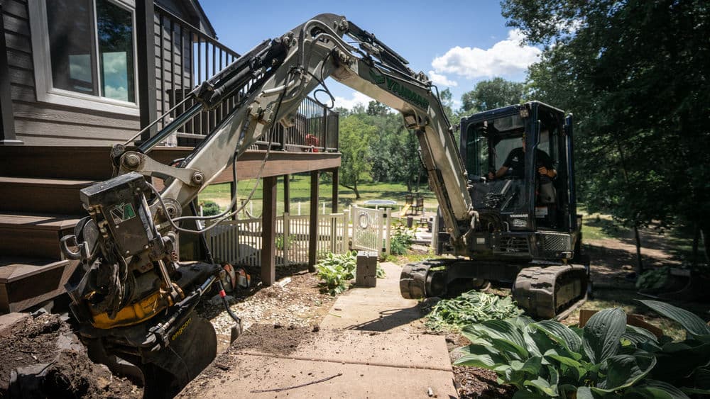 Excavator working near a house, with green landscaping and trees in the background.