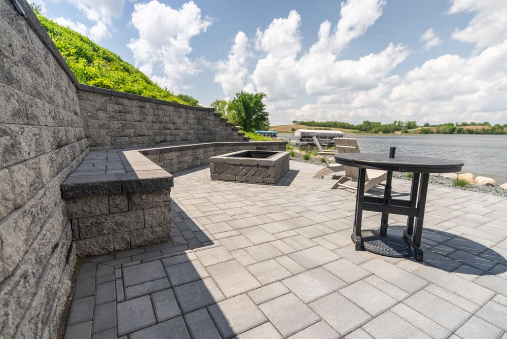Patio by the river with stone seating, fire pit, and a table under a blue sky.