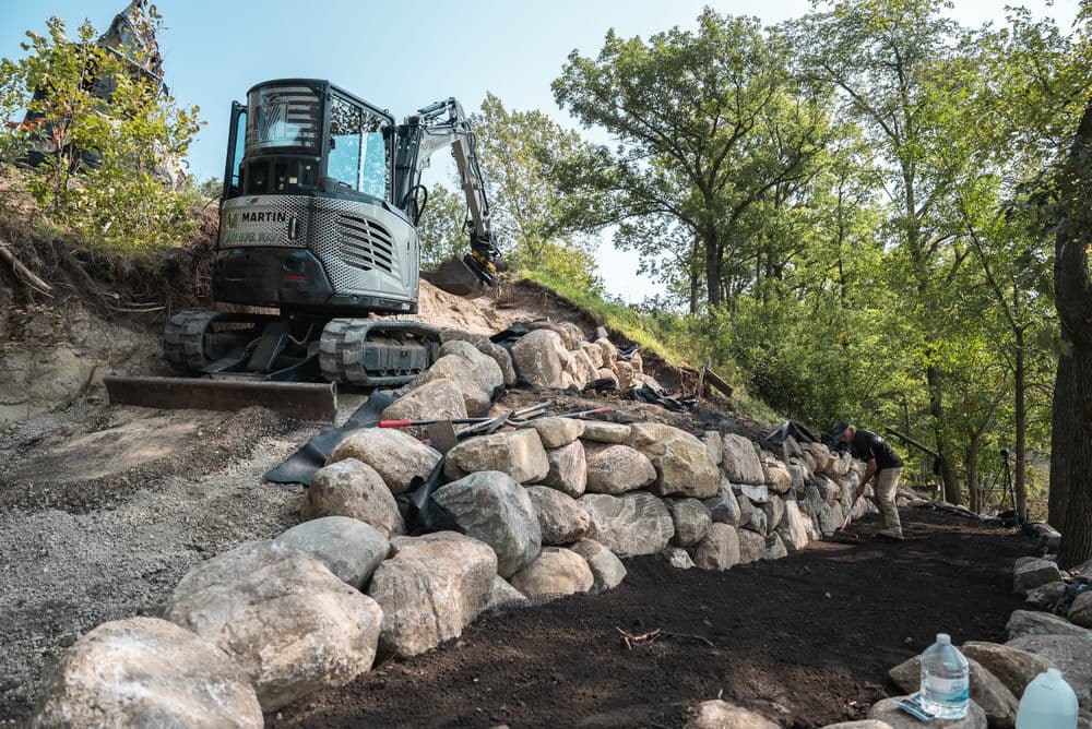 Excavator working on a stone retaining wall with a worker landscaping nearby.