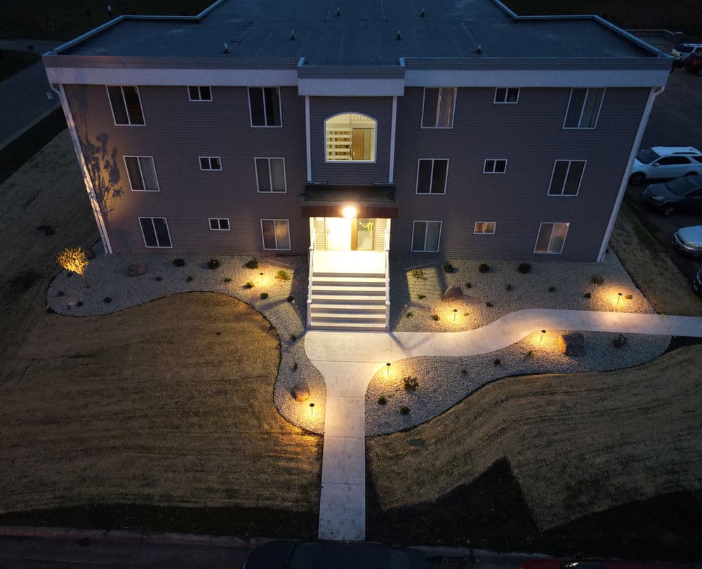 Aerial view of a well-lit apartment building entrance with landscaped grounds at night.