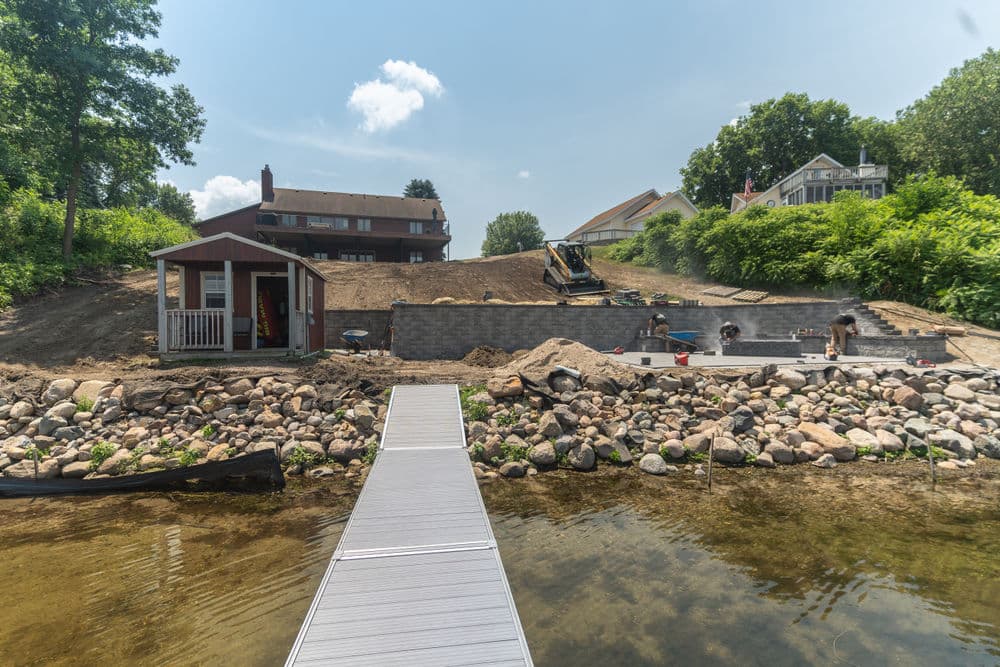Construction site by a lake featuring a wooden dock, equipment, and a landscaped yard.