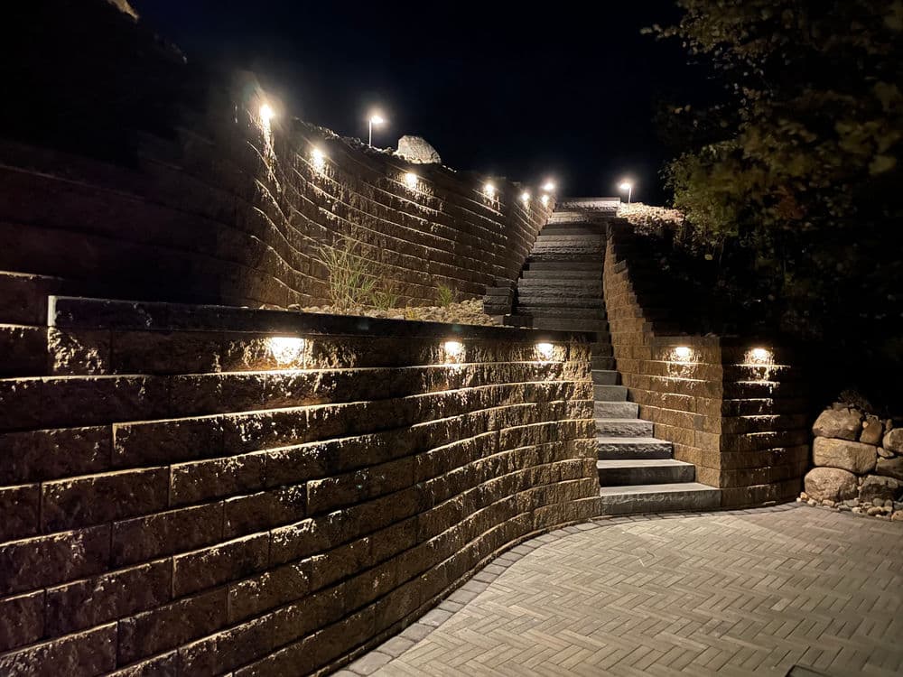 Illuminated stone staircase at night with landscape lighting and textured brick pathway.