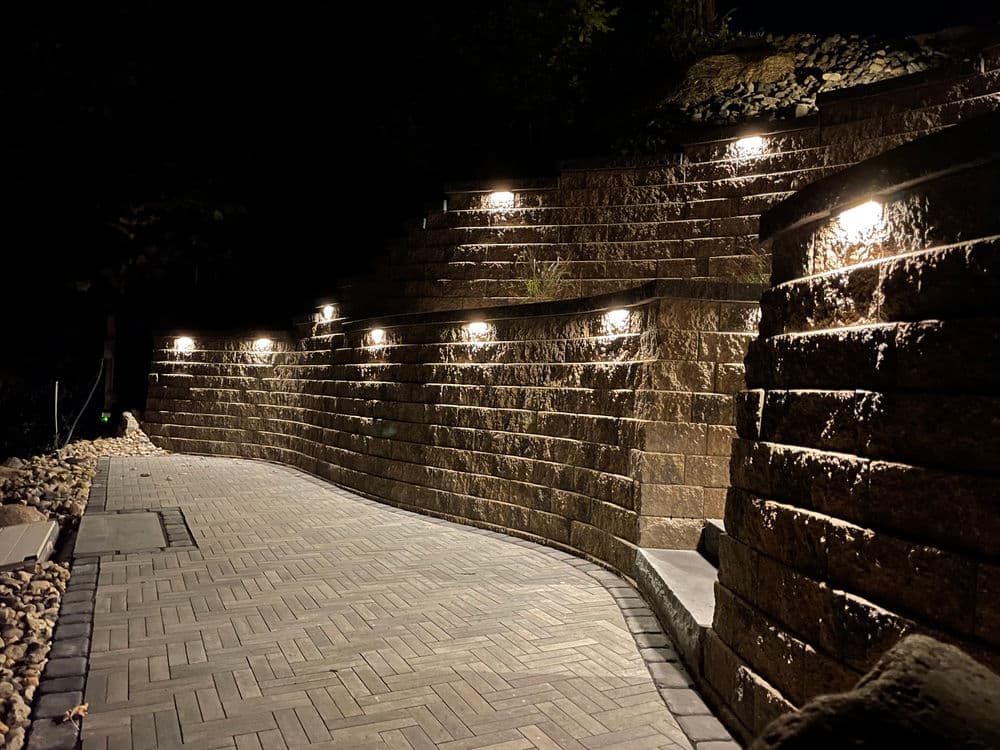 Illuminated stone retaining wall and pathway at night with landscaping lights.
