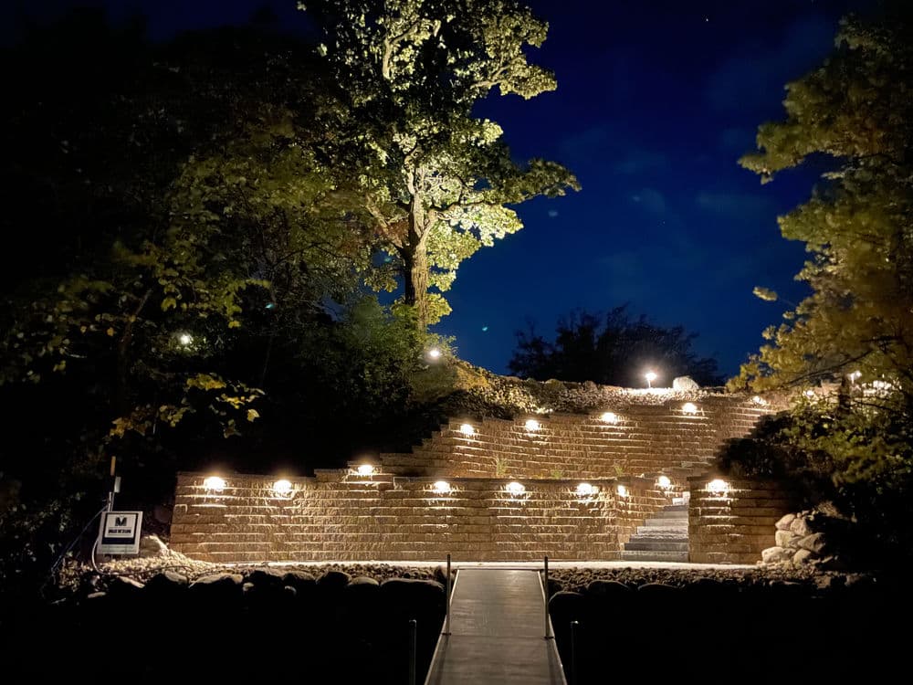 Illuminated outdoor amphitheater at night surrounded by trees and landscaped rocky area.