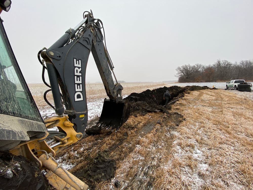 Excavator digging a trench in a snowy field, with a worker and truck nearby.