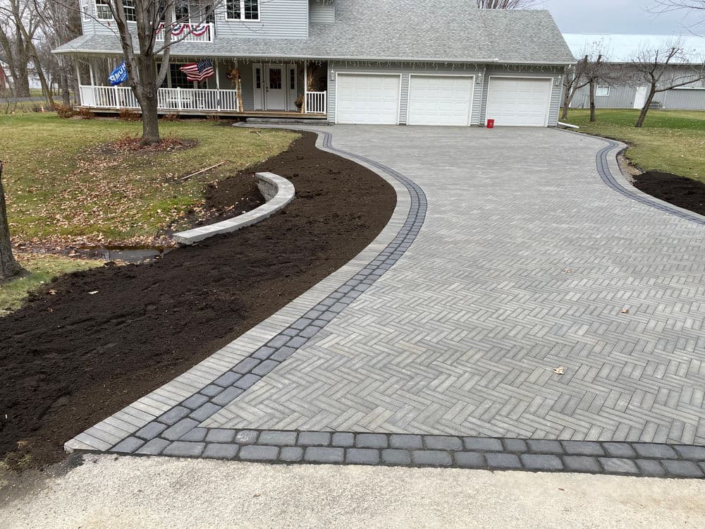 Paved driveway with herringbone pattern leading to a house with garage and landscaped yard.