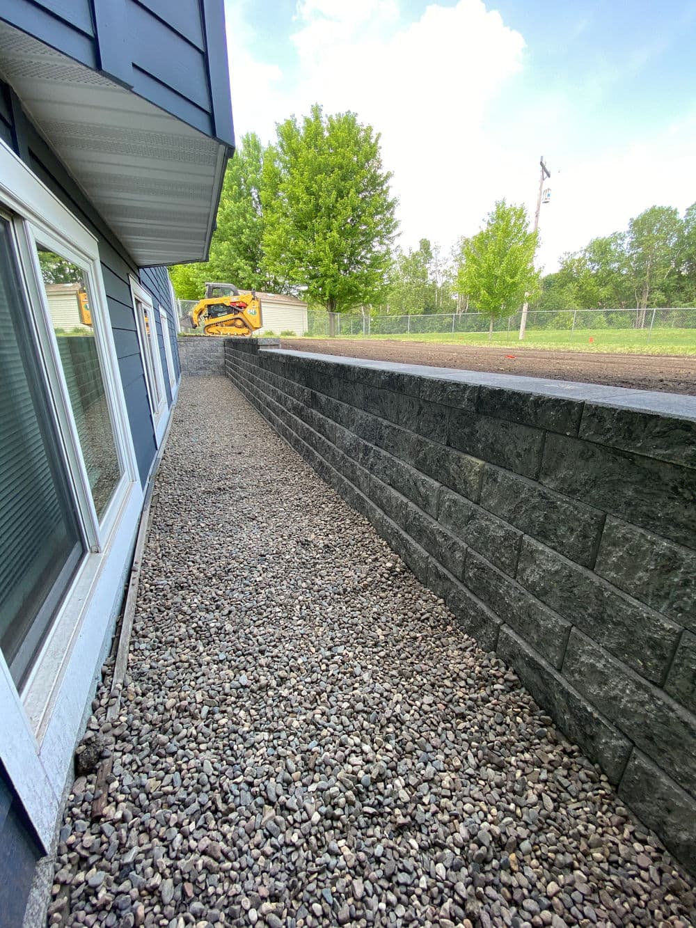 Retaining wall made of dark stone, lined with gravel, alongside a blue house and green trees.