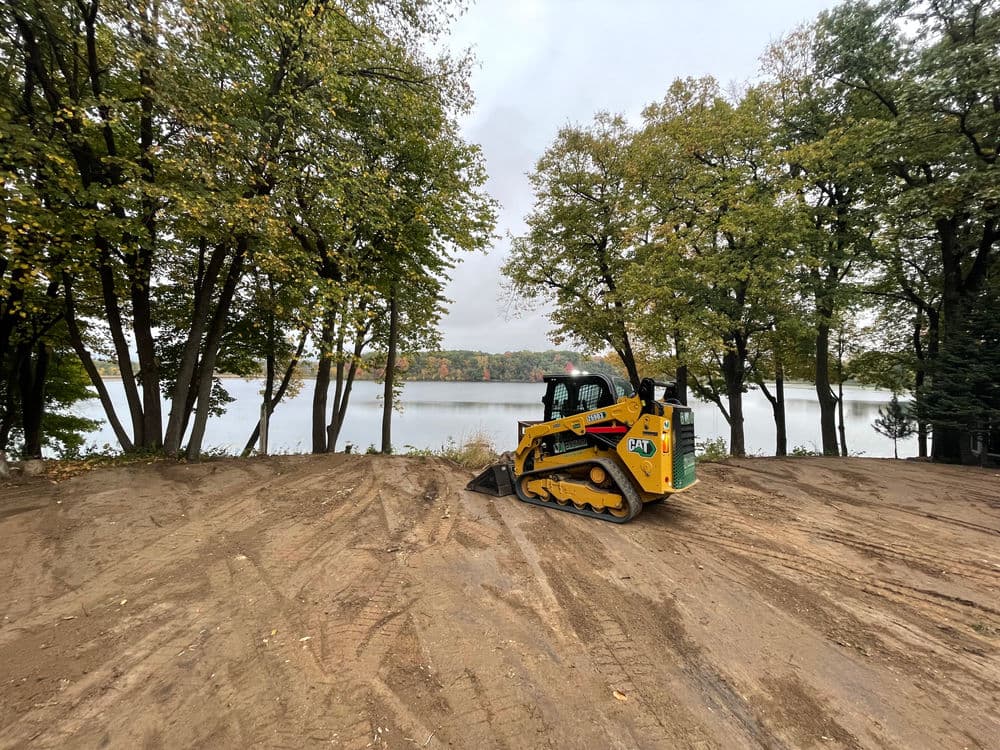 Caterpillar skid steer loader on cleared dirt next to a lake surrounded by trees.