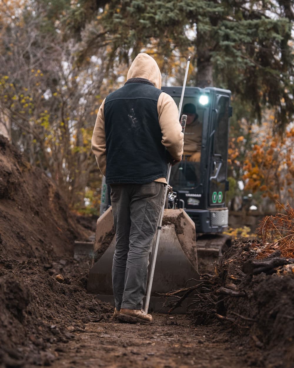 Construction worker with a surveying stick walking towards an excavator in a muddy trench.