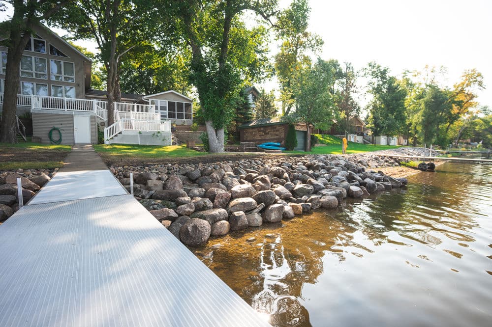 Lakefront property with a dock, rocky shoreline, and lush green trees in a sunny setting.
