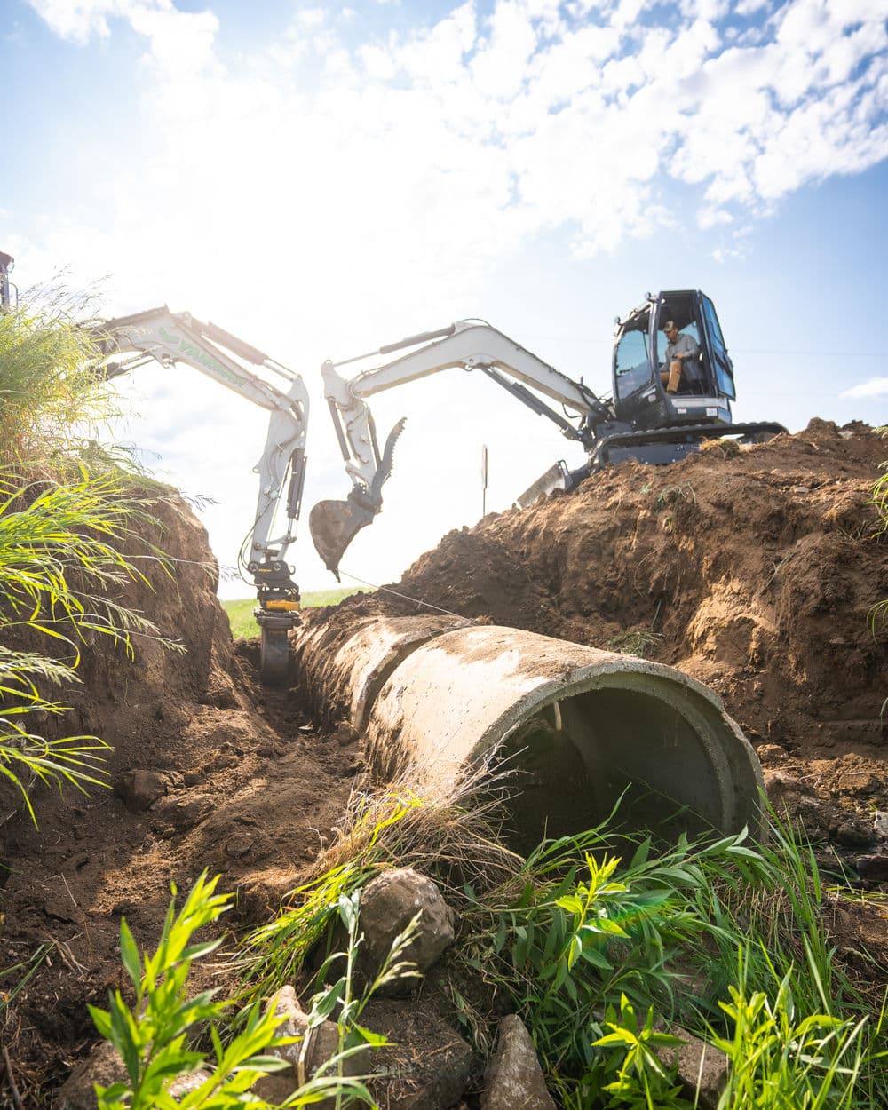 Excavator digging near a concrete pipe under a bright sky and lush greenery.