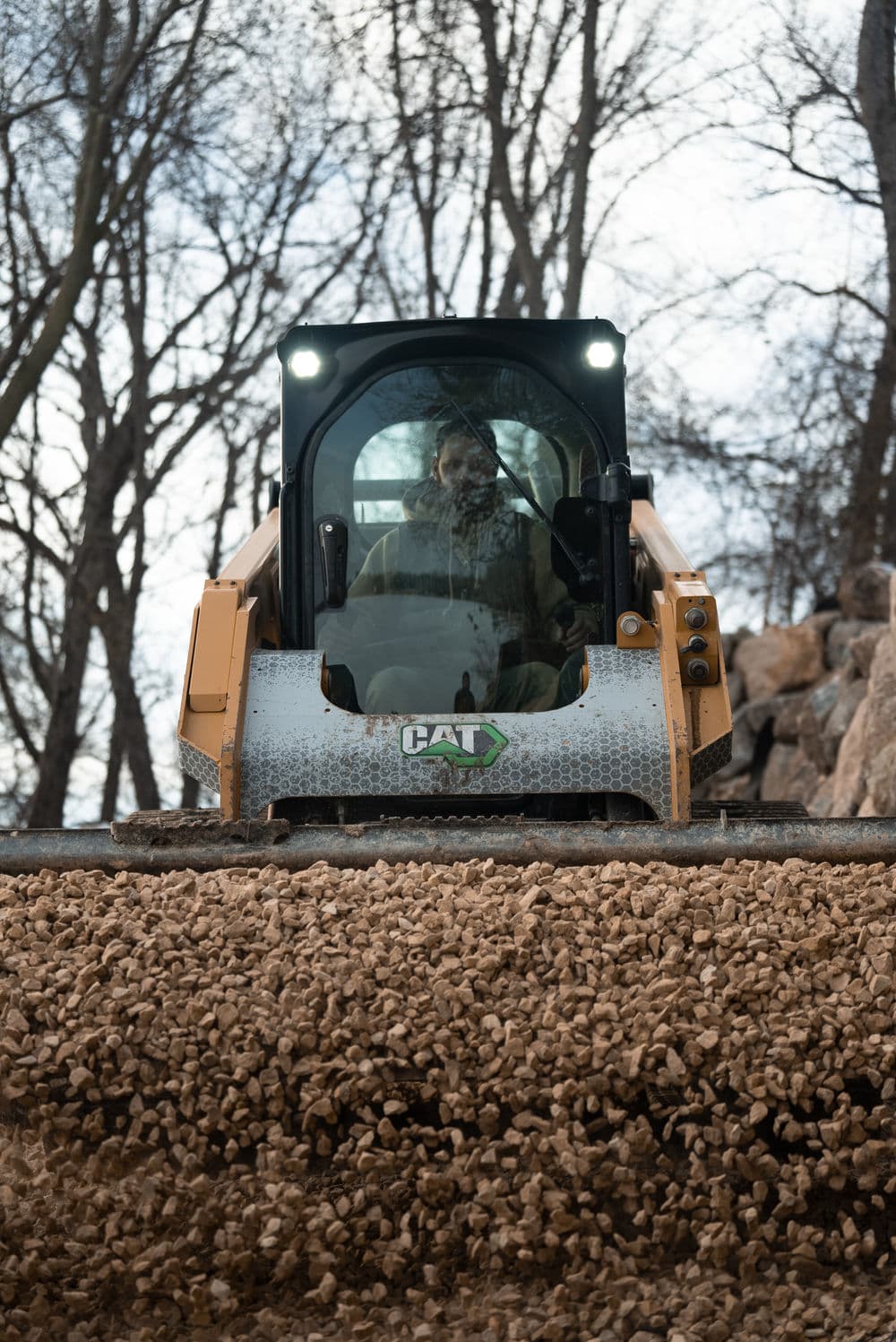 CAT skid steer loader operating on rocky terrain with trees in the background.