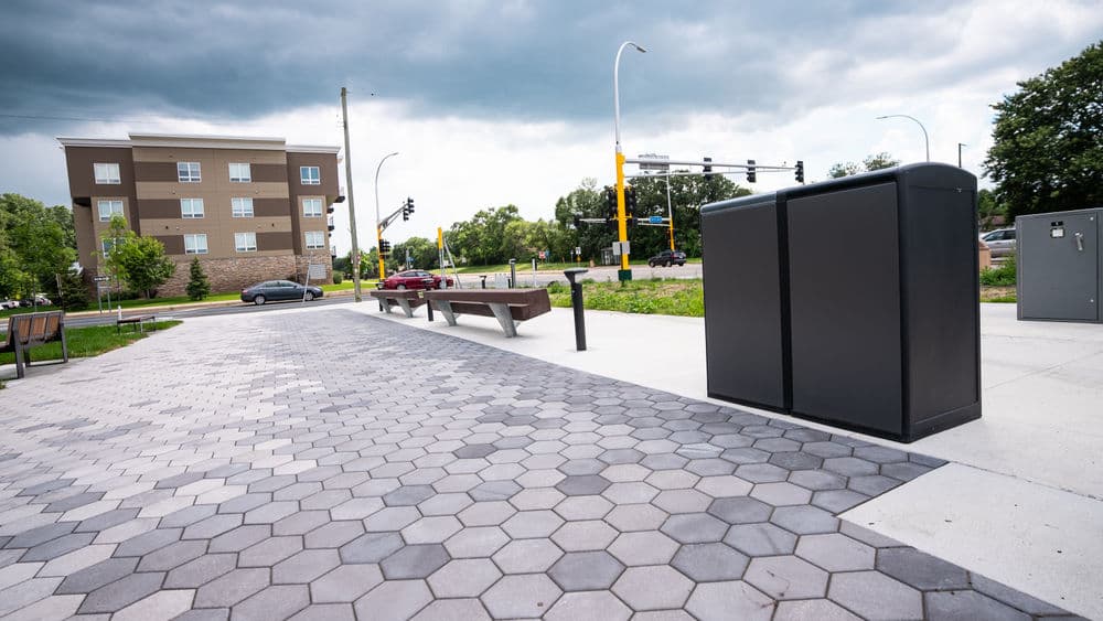 Urban park featuring hexagonal paving, benches, and a utility box under a cloudy sky.