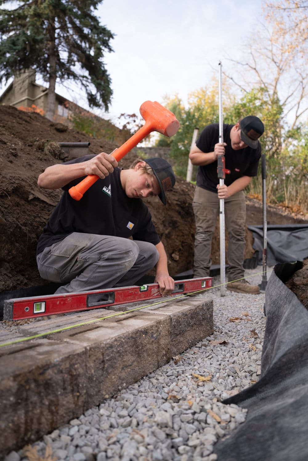 Two workers leveling gravel with a hammer and measuring tool during a landscaping project.