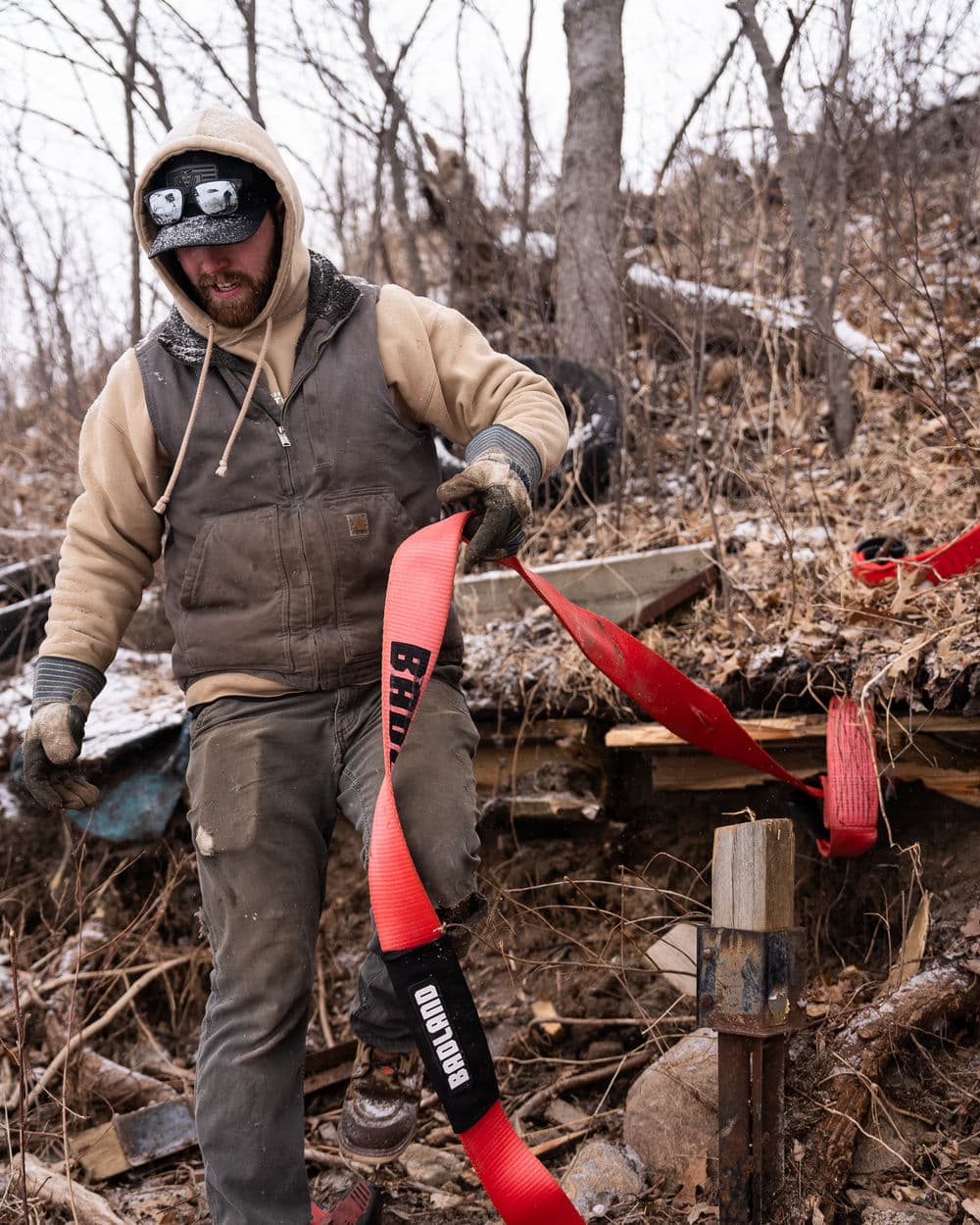 Man in a hooded jacket adjusting red straps in a wooded area with snow on the ground.