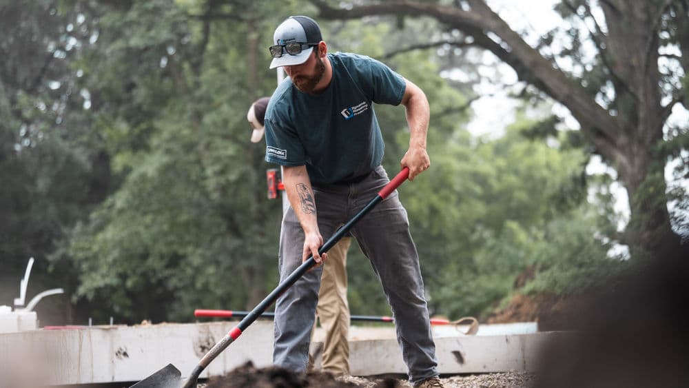 Construction worker using a shovel on a job site surrounded by trees and equipment.