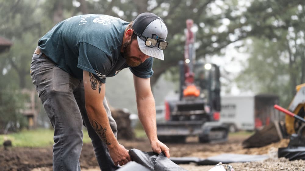 Man working on a construction site, laying down black landscaping fabric. Heavy machinery in background.