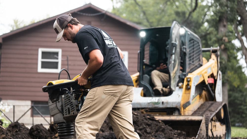 Construction worker operating a compactor near a CAT skid steer loader at a home site.