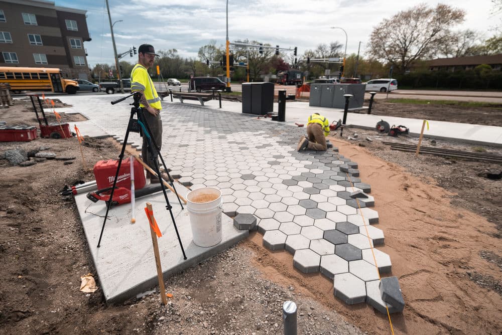 Construction workers install hexagonal pavers on a sidewalk, with tools and machinery nearby.