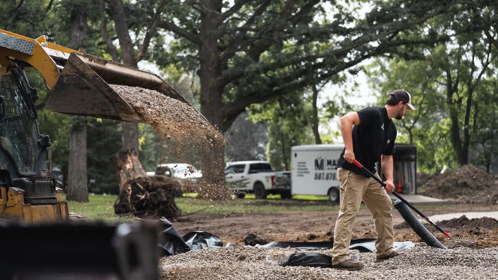 Worker operating a loader to pour gravel on a landscaping site. Trees in the background.