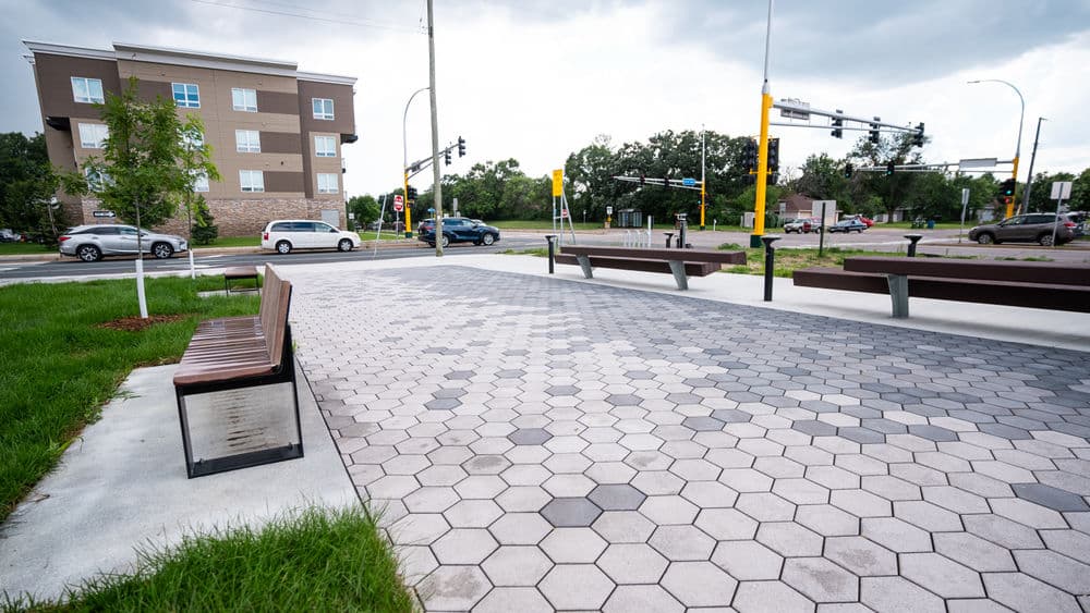Modern urban plaza with hexagonal pavers, benches, and green space near a traffic intersection.