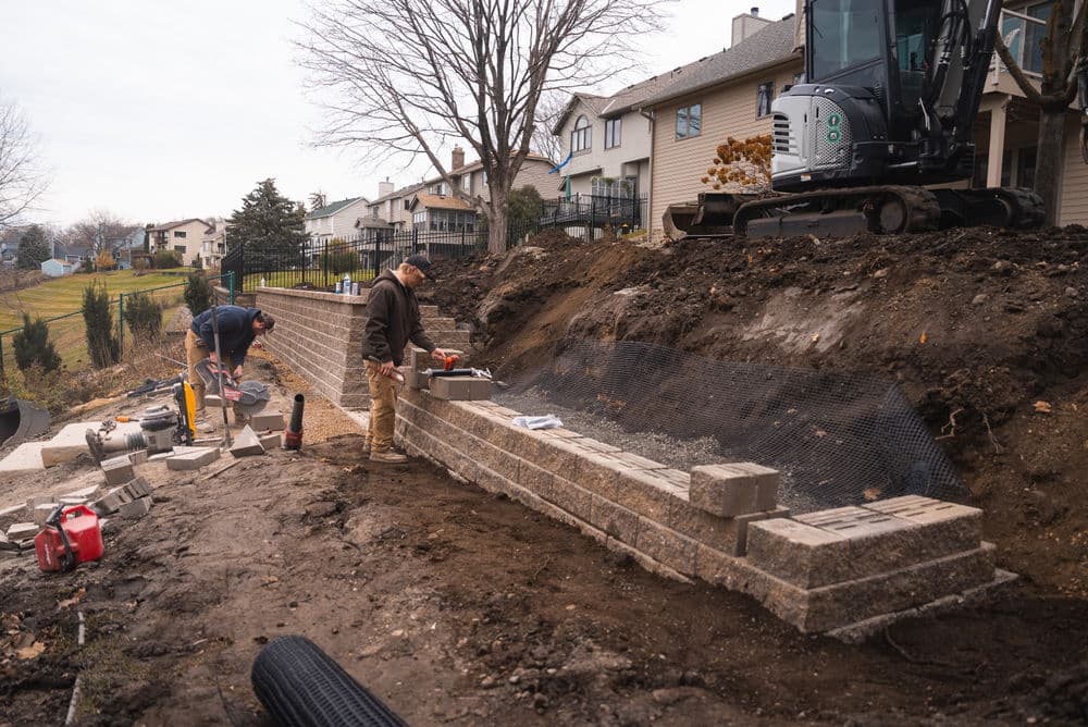 Two workers constructing a stone retaining wall in a residential yard with machinery nearby.