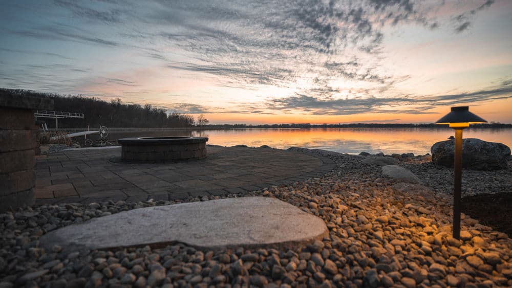 Sunset over a serene lake with a stone patio and warm outdoor lighting.