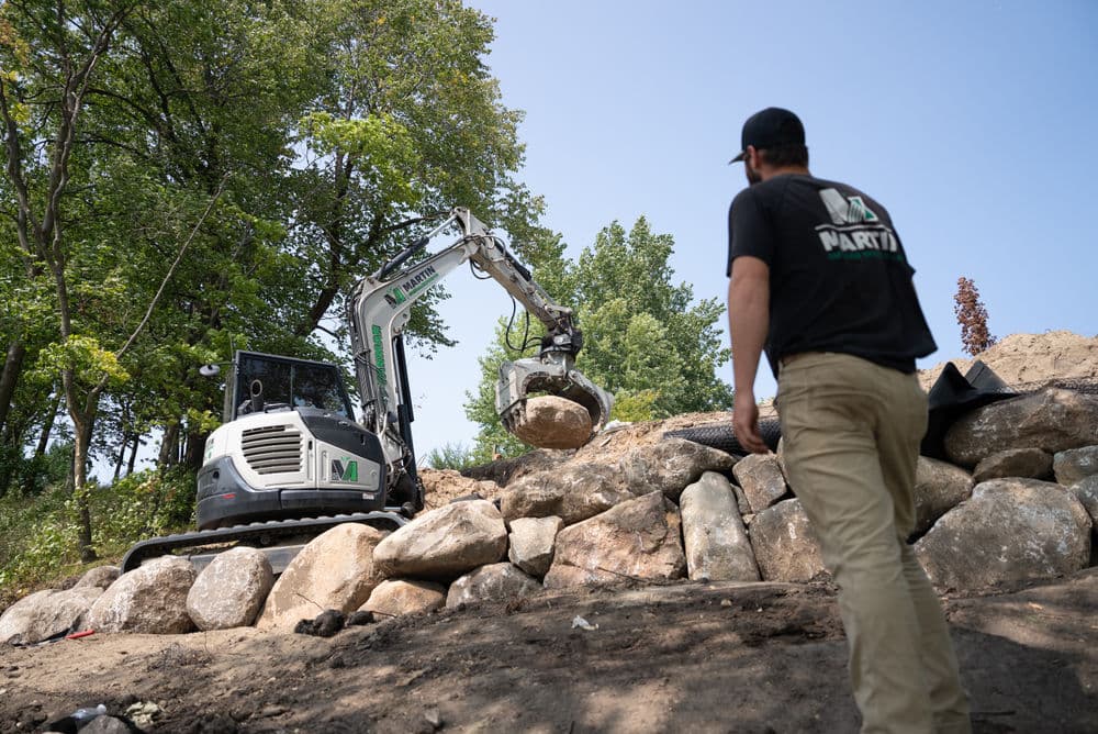 Excavator maneuvering large rock at a construction site with worker observing.
