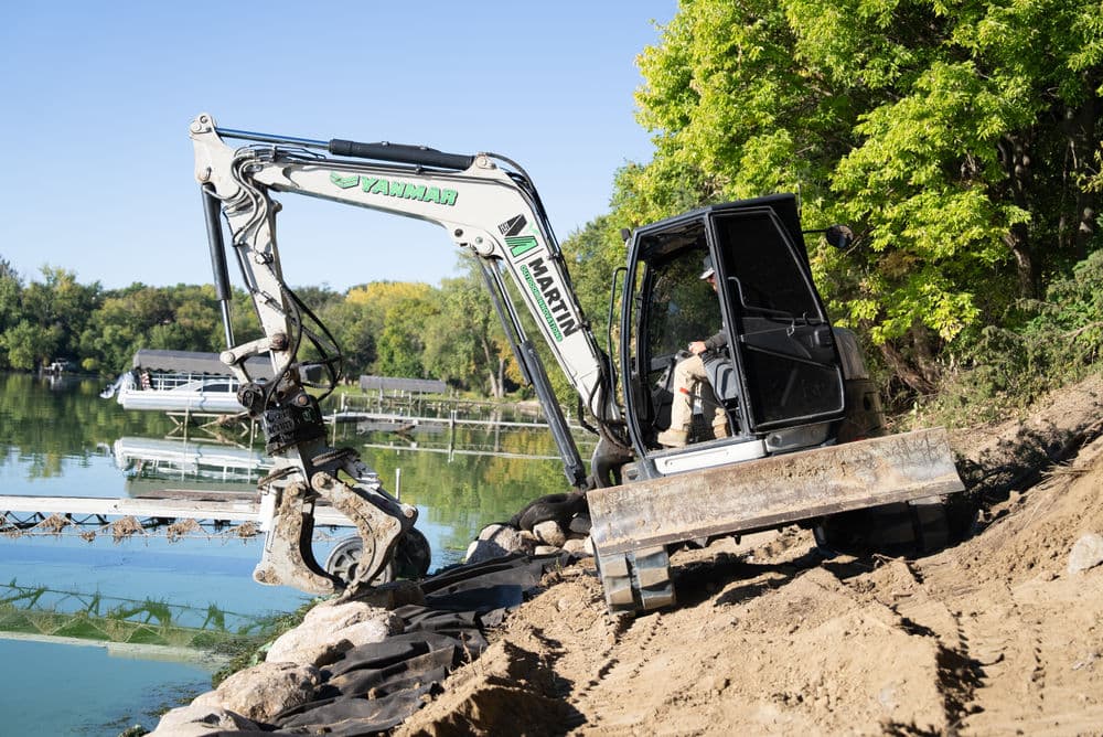 Excavator on slope near a lake, preparing land for construction with trees in the background.