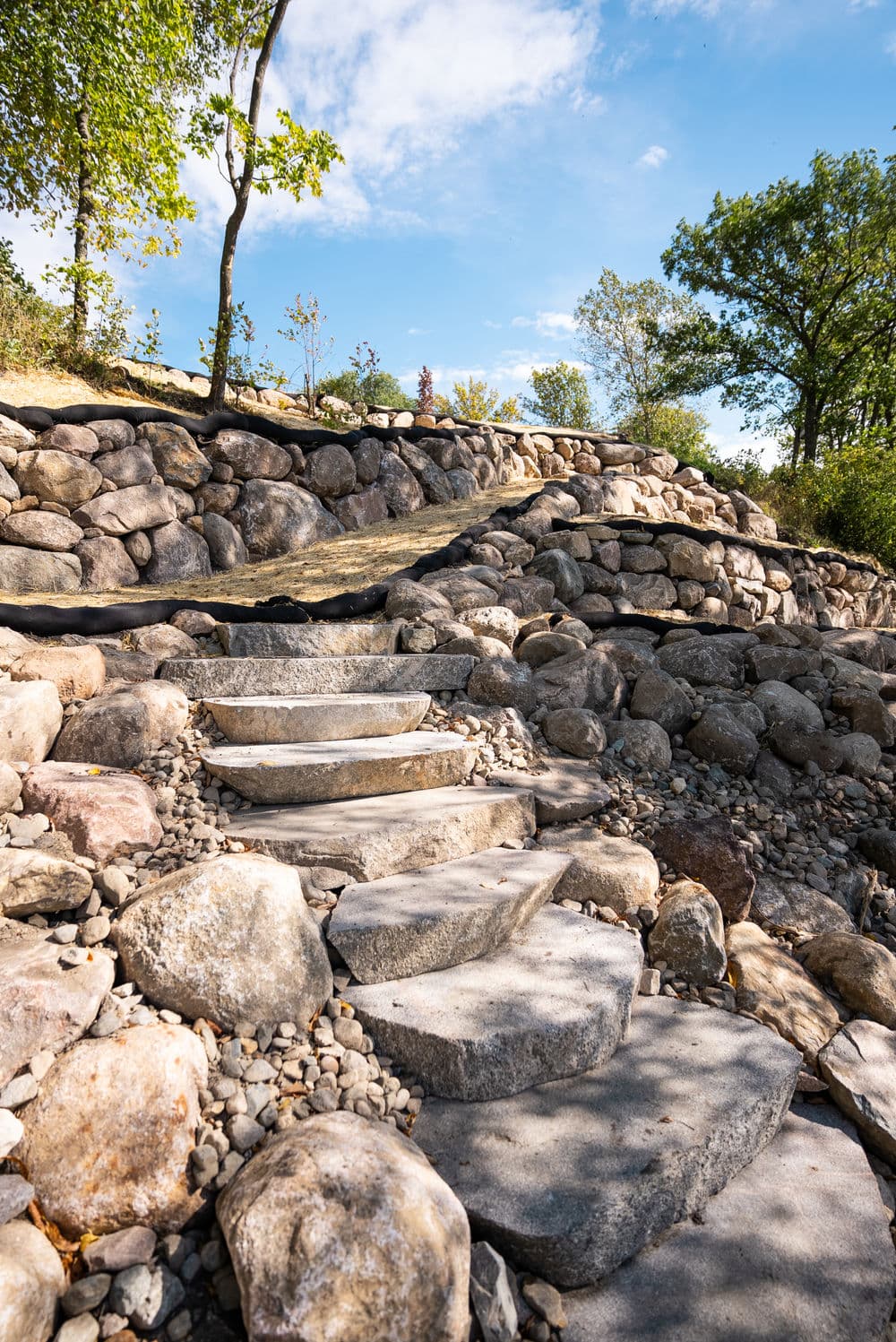 Stone steps lead up a rocky pathway surrounded by trees and blue sky.