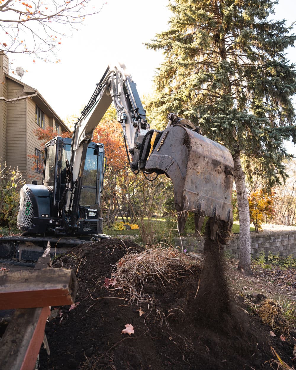 Excavator digging in a backyard, lifting soil and debris with trees in the background.