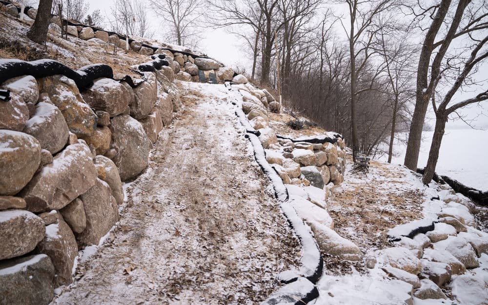 Snow-covered dirt path alongside stone wall in a winter landscape with bare trees.