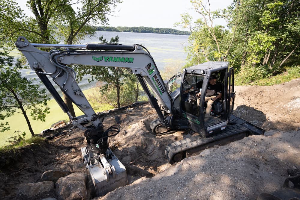 Excavator operating on a hillside by a lake, surrounded by trees and green vegetation.