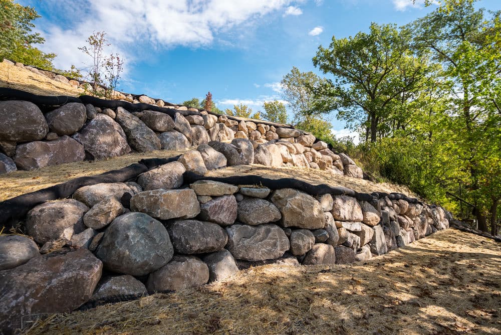 Stone retaining wall on a hillside with greenery and blue sky in the background.