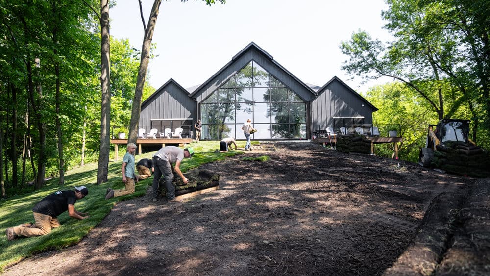 Workers installing sod on a hillside near a modern glass house surrounded by trees.