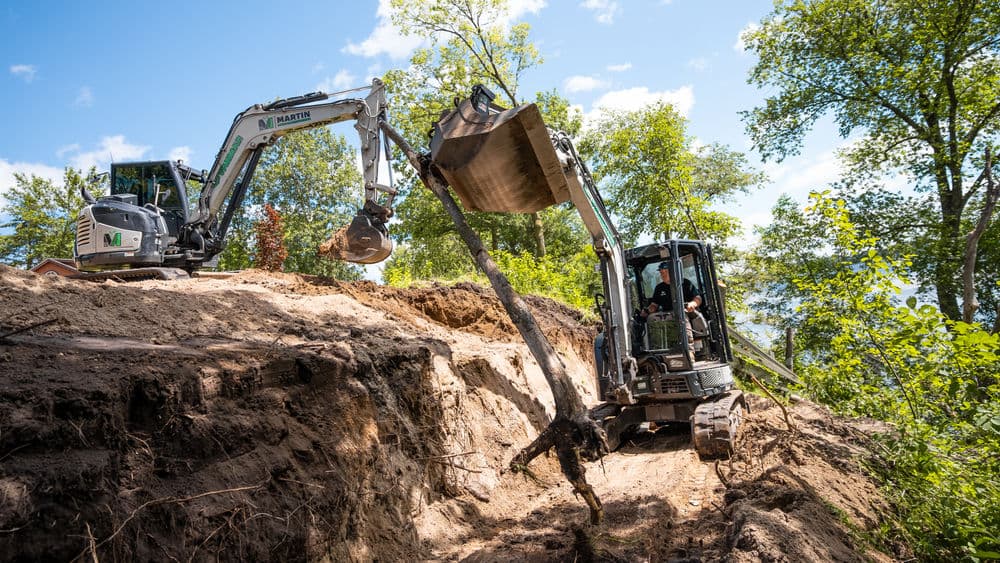 Excavators working on a hillside excavation project with trees and blue sky in the background.