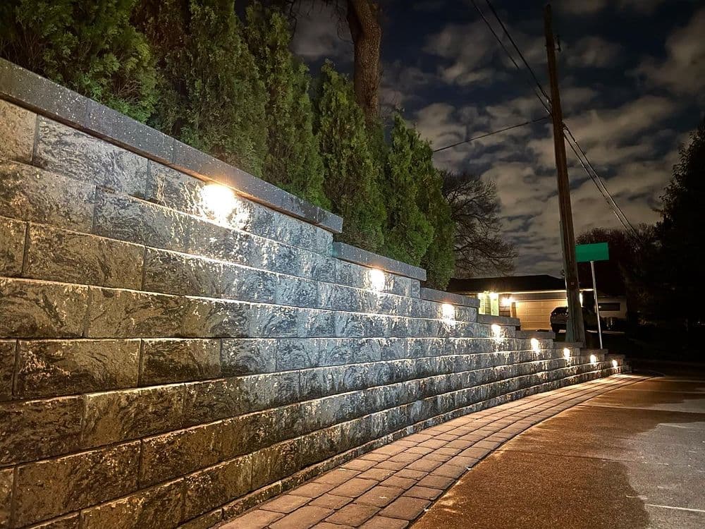 Illuminated stone wall at night with landscaped greenery and cloudy sky backdrop.