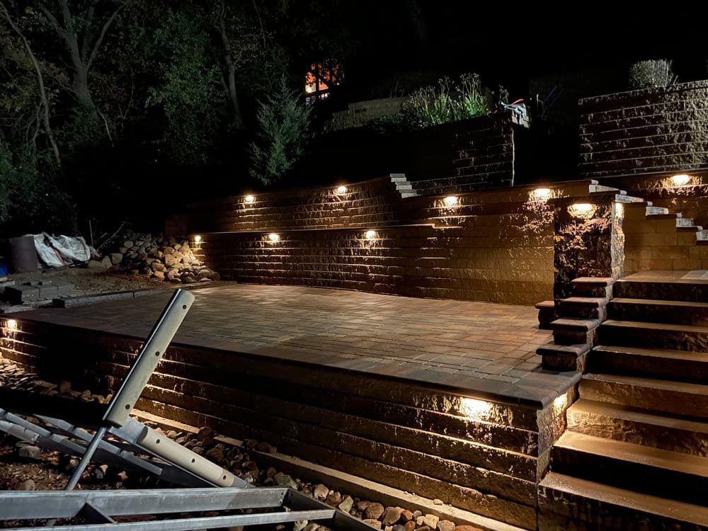 Illuminated outdoor patio at night featuring stone steps and walls with landscaping.