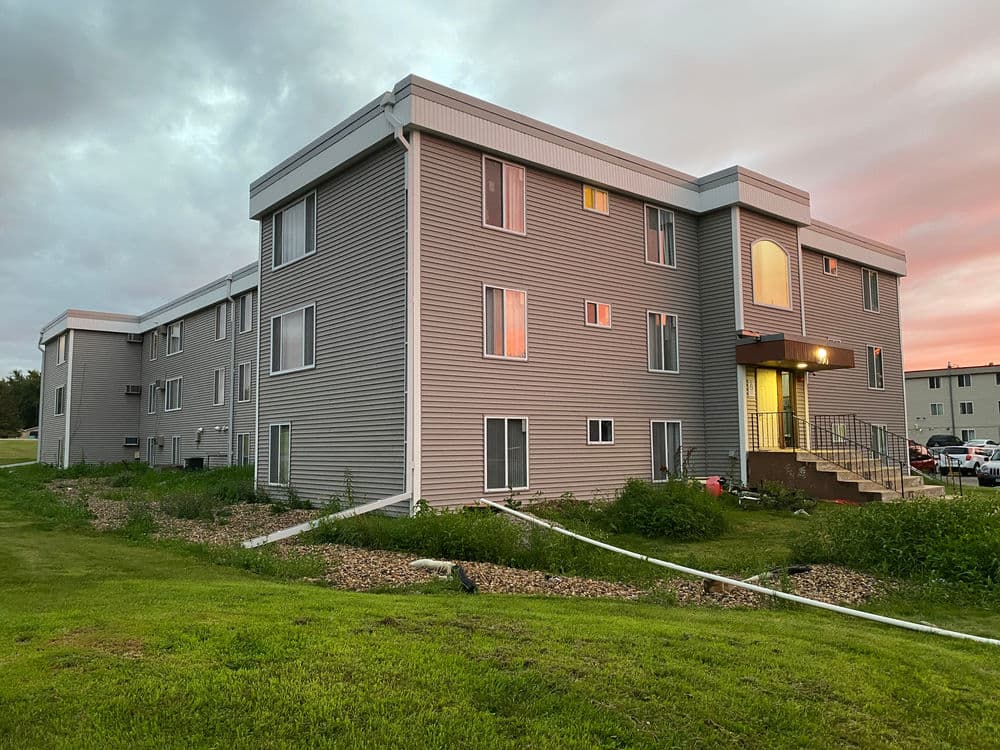 Modern apartment building with multiple windows and stairs, surrounded by green lawn at sunset.