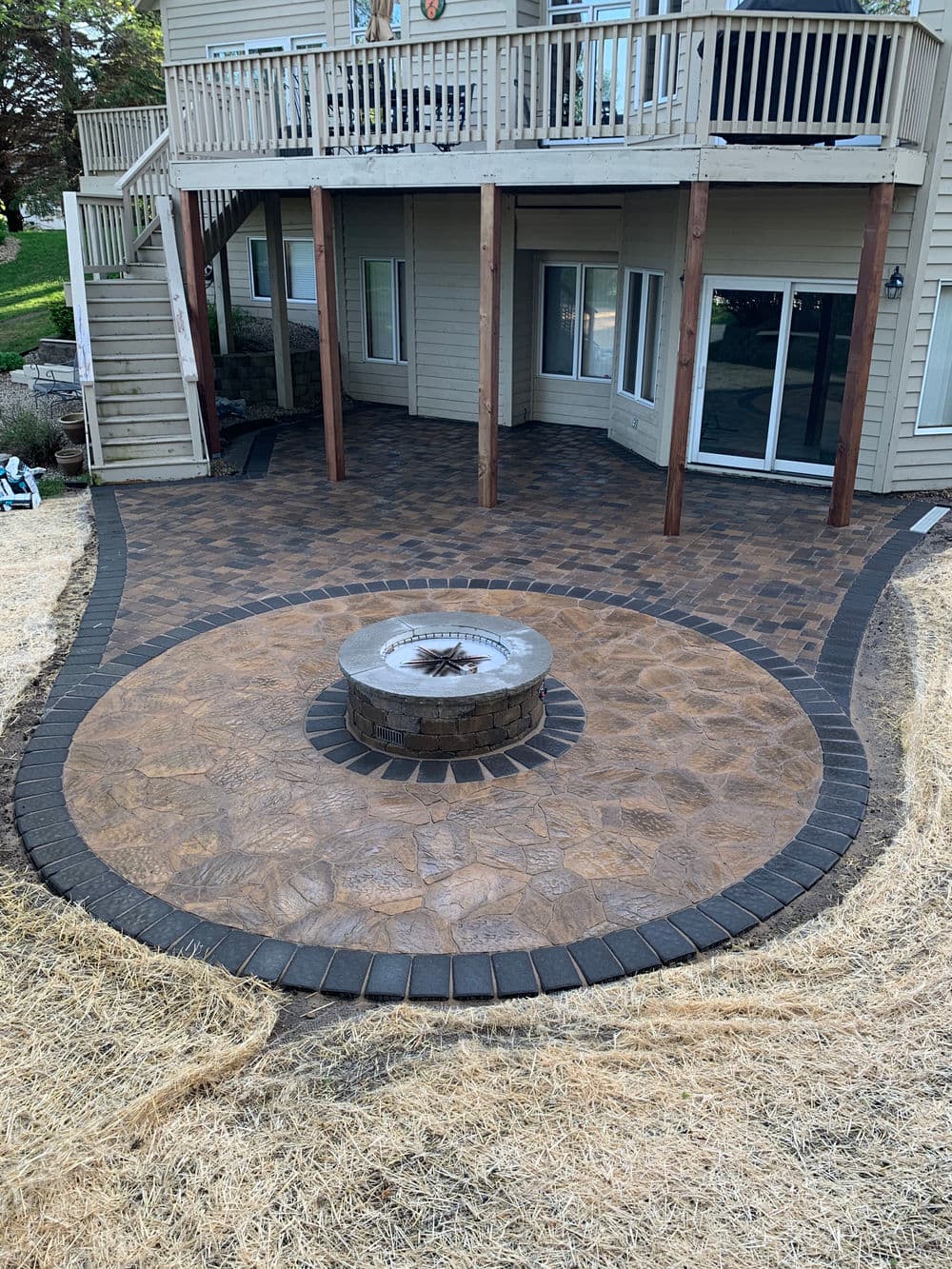 Patio with circular stone fire pit and decorative paving at a home with a wooden deck.