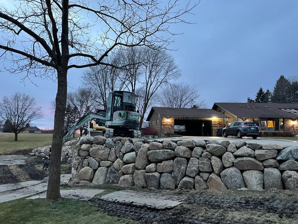 Excavator working on a stone wall at dusk with homes in the background.