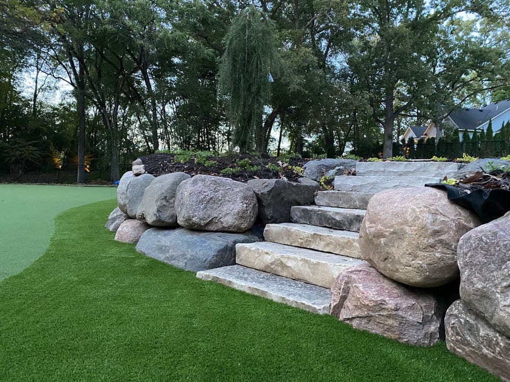 Stone steps with large rocks in a landscaped yard, surrounded by greenery and trees.