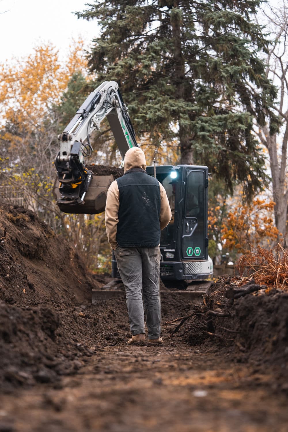 Construction worker operating a mini excavator in a trench surrounded by autumn trees.