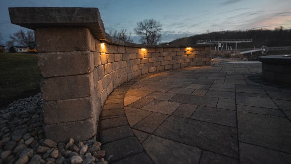 Curved stone wall with ambient lighting at dusk near a lake shore.
