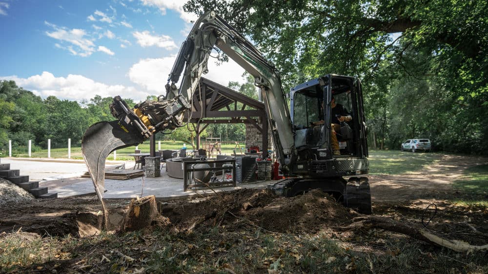 Excavator digging near a construction site under clear blue skies and trees.