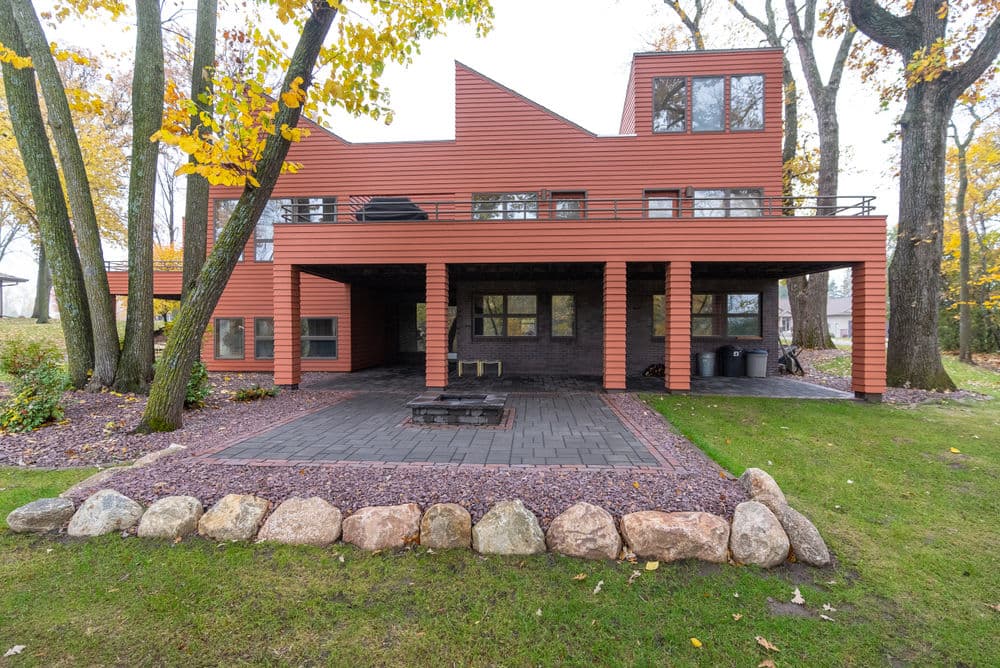 Modern home with orange siding, large windows, and a patio surrounded by trees and stone landscaping.