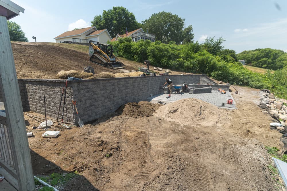 Construction of a stone retaining wall with workers and machinery in a landscaped area.